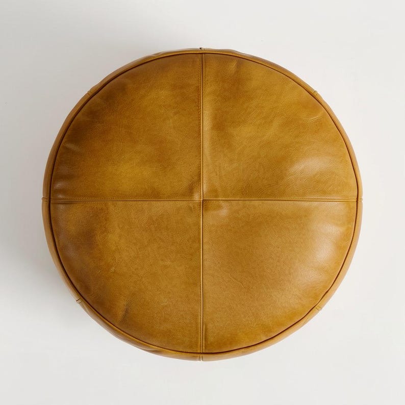 May include: A round, tan leather ottoman. The top is divided into four sections by stitched seams. The ottoman has a smooth, slightly textured surface and a rounded edge.