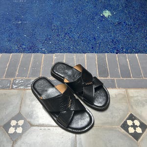 May include: A pair of black leather sandals with a crisscross design. The sandals have a textured appearance and are placed on a tiled surface. The background features a blue pool.