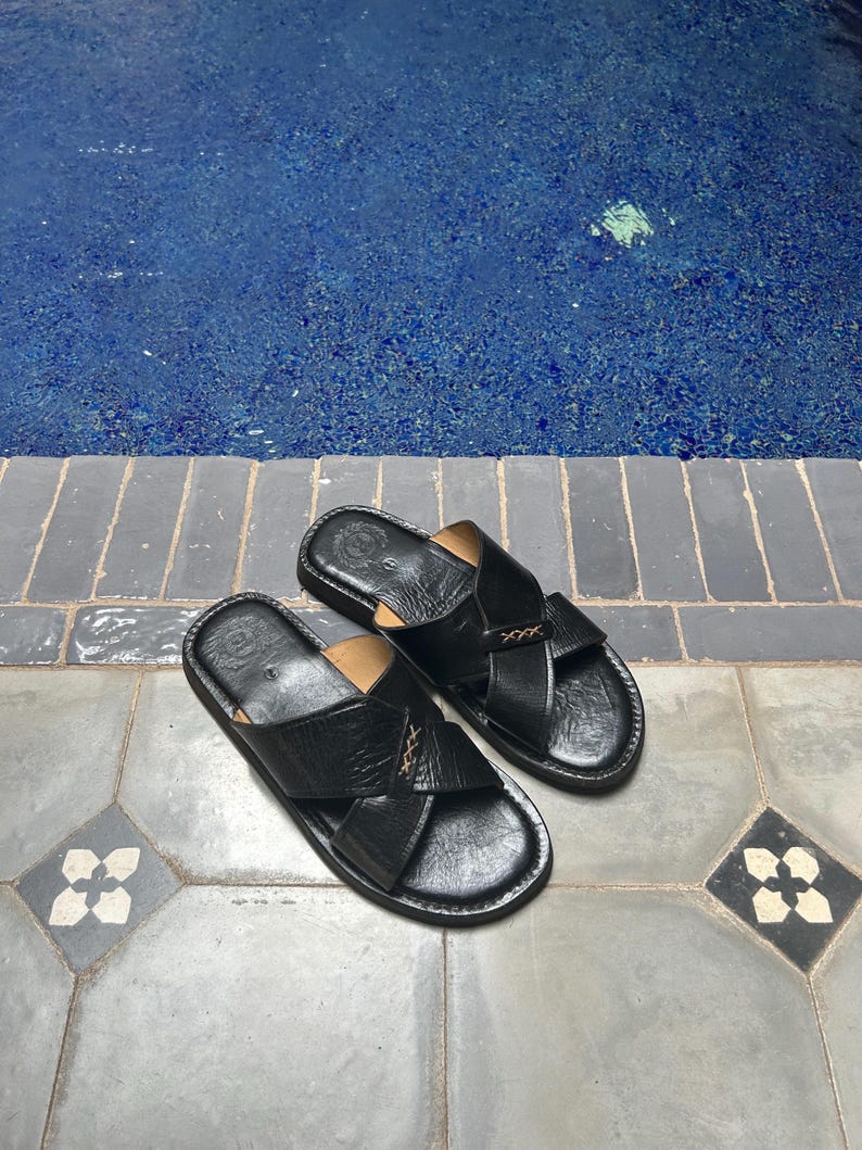 May include: A pair of black leather sandals with a crisscross design. The sandals have a textured appearance and are placed on a tiled surface. The background features a blue pool.