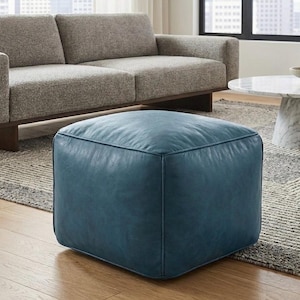 May include: A square, teal leather ottoman. The ottoman is placed on a patterned rug in front of a light gray sofa. The ottoman is a cube shape and appears to be made of soft leather.