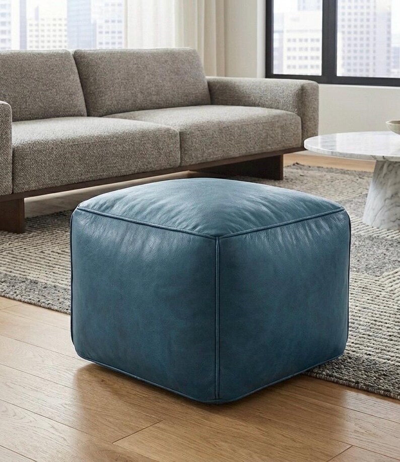 May include: A square, teal leather ottoman. The ottoman is placed on a patterned rug in front of a light gray sofa. The ottoman is a cube shape and appears to be made of soft leather.