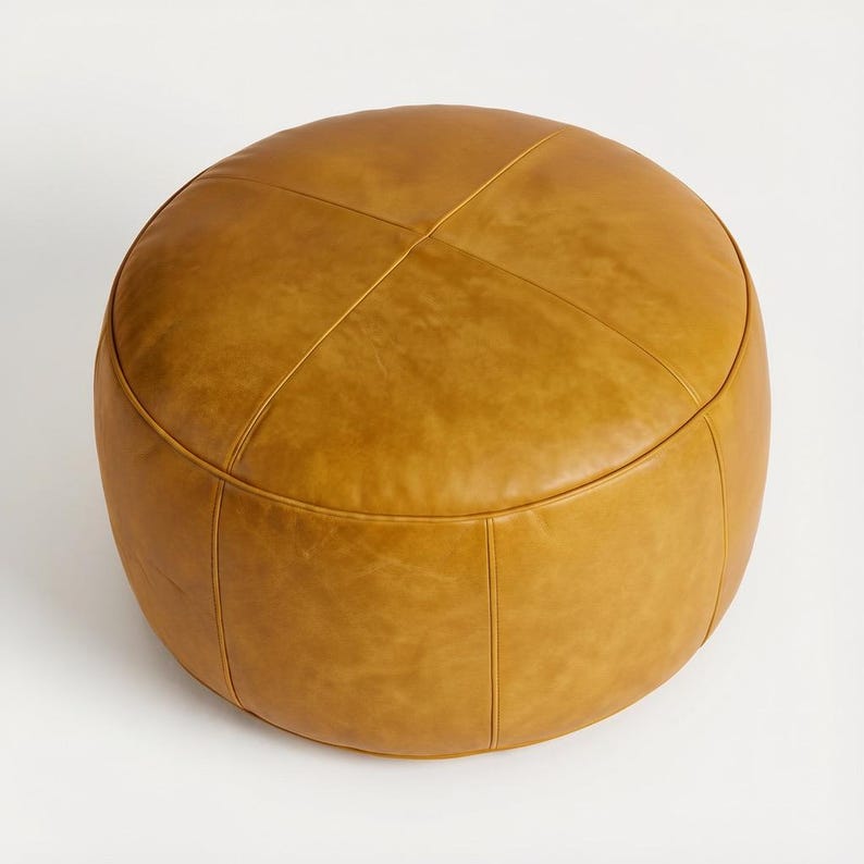 May include: A round, mustard-colored ottoman with a stitched, cross-patterned top. The leather-like material has a slight sheen, and the seams are visible. This footstool is a stylish addition to any living space.