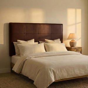 May include: A bedroom scene featuring a bed with a brown leather headboard and beige bedding. The bed is made with several pillows and a duvet. A wooden nightstand with a lamp sits beside the bed, and the room has a warm, inviting atmosphere.