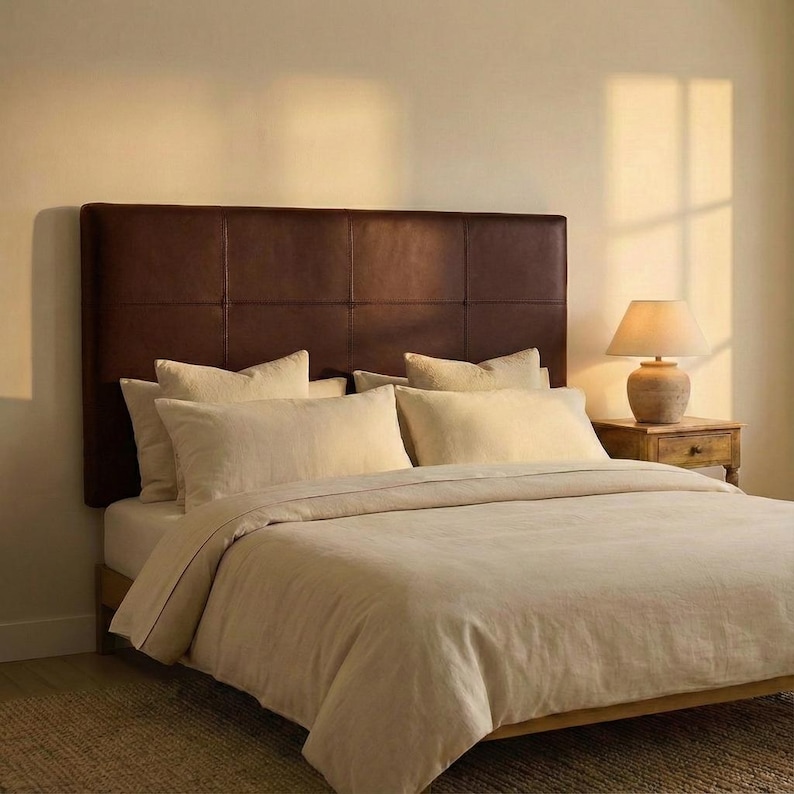 May include: A bedroom scene featuring a bed with a brown leather headboard and beige bedding. The bed is made with several pillows and a duvet. A wooden nightstand with a lamp sits beside the bed, and the room has a warm, inviting atmosphere.