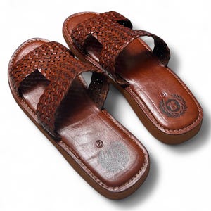 May include: A pair of brown leather sandals with a woven strap design. The sandals have a dark brown sole and a stitched border. The footbed has a logo and the number 2 is visible.