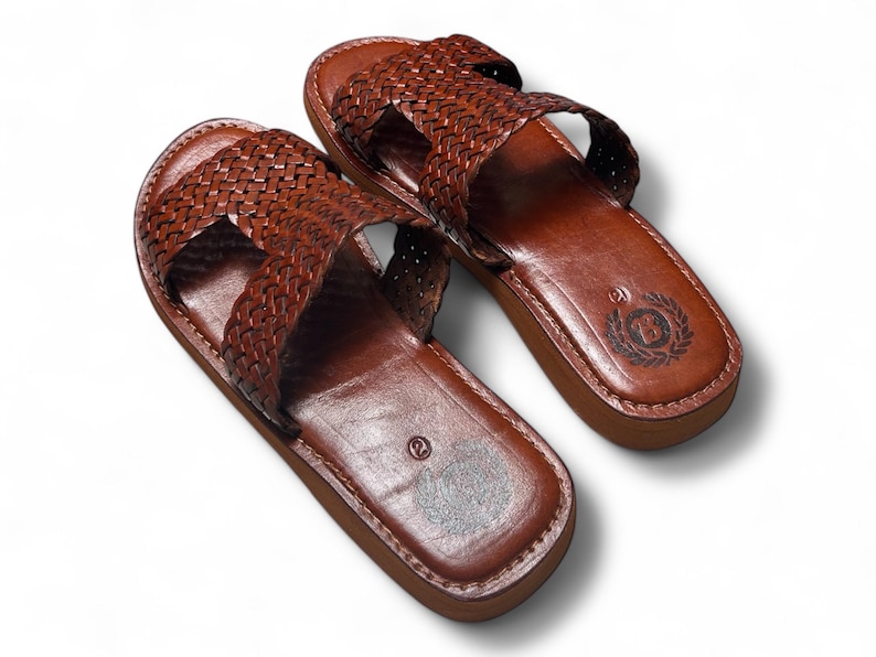 May include: A pair of brown leather sandals with a woven strap design. The sandals have a dark brown sole and a stitched border. The footbed has a logo and the number 2 is visible.