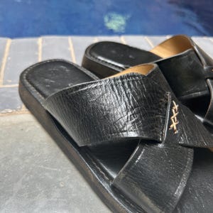 May include: Black leather sandals with a crisscross strap design. The sandals have a textured appearance and a dark sole. The background shows a blue surface, possibly a pool.