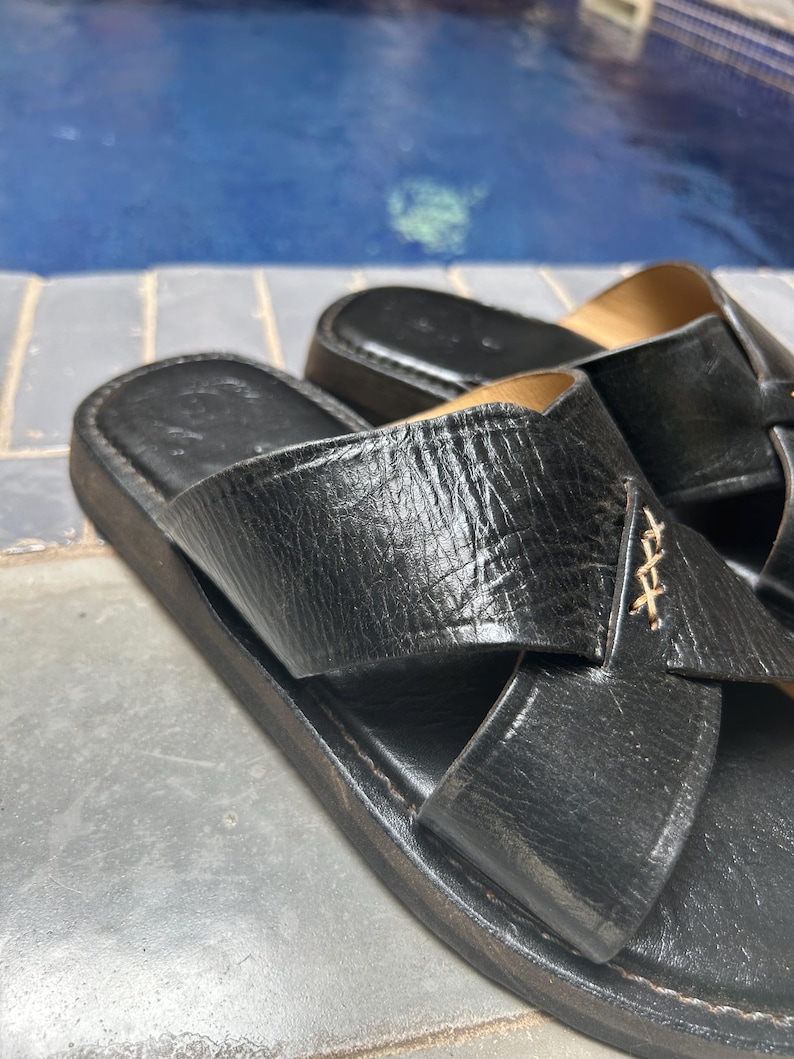 May include: Black leather sandals with a crisscross strap design. The sandals have a textured appearance and a dark sole. The background shows a blue surface, possibly a pool.