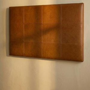 May include: A rectangular, brown leather headboard with a grid pattern of stitched squares. The headboard is mounted on a light-colored wall, and the leather has a warm, inviting tone. The headboard is a stylish addition to any bedroom.