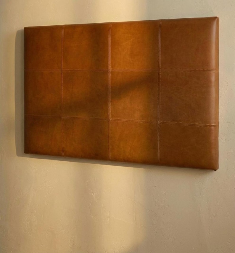 May include: A rectangular, brown leather headboard with a grid pattern of stitched squares. The headboard is mounted on a light-colored wall, and the leather has a warm, inviting tone. The headboard is a stylish addition to any bedroom.