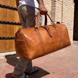 May include: A large, tan leather duffel bag with a cylindrical shape. The bag has two top handles, a shoulder strap, and a side zipper pocket. The bag is carried by a person. The bag is made of leather and has a buckle closure.