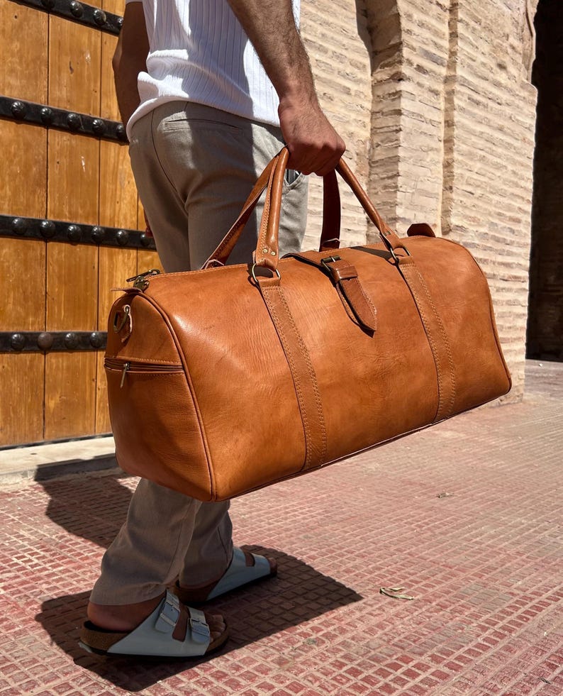May include: A large, tan leather duffel bag with a cylindrical shape. The bag has two top handles, a shoulder strap, and a side zipper pocket. The bag is carried by a person. The bag is made of leather and has a buckle closure.