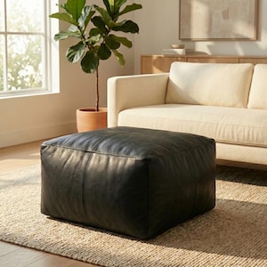 Peut inclure: Un pouf carré en cuir noir. Il est placé sur un tapis tissé devant un canapé crème. Un pot de plante est visible en arrière-plan, près d'une fenêtre. Le pouf est un meuble polyvalent.