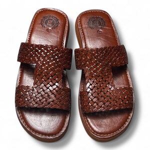 May include: A pair of brown leather sandals with a woven strap design. The sandals have a rounded toe and a thick sole. The top of the sandal features a circular emblem. The sandals are displayed on a white background.