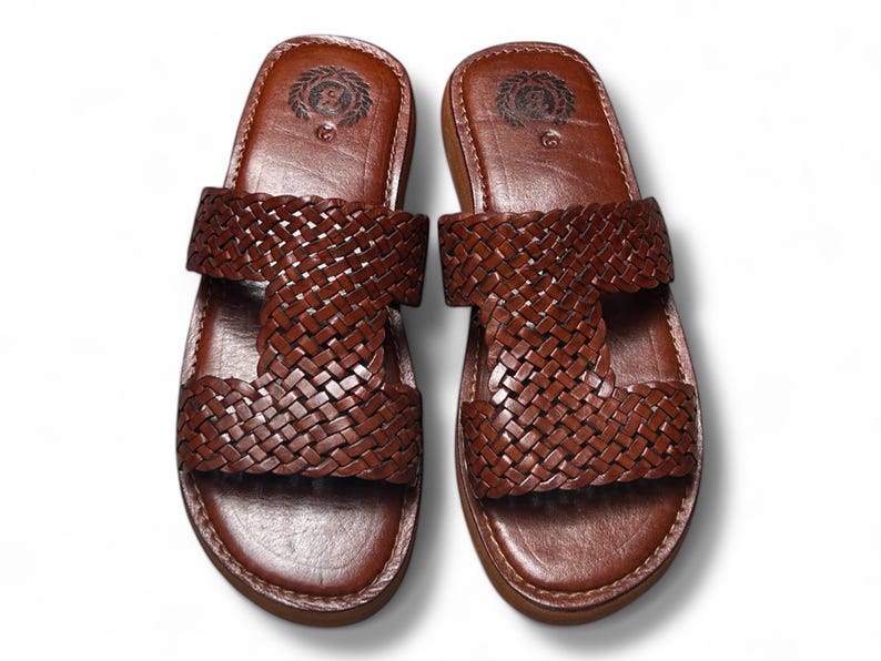 May include: A pair of brown leather sandals with a woven strap design. The sandals have a rounded toe and a thick sole. The top of the sandal features a circular emblem. The sandals are displayed on a white background.