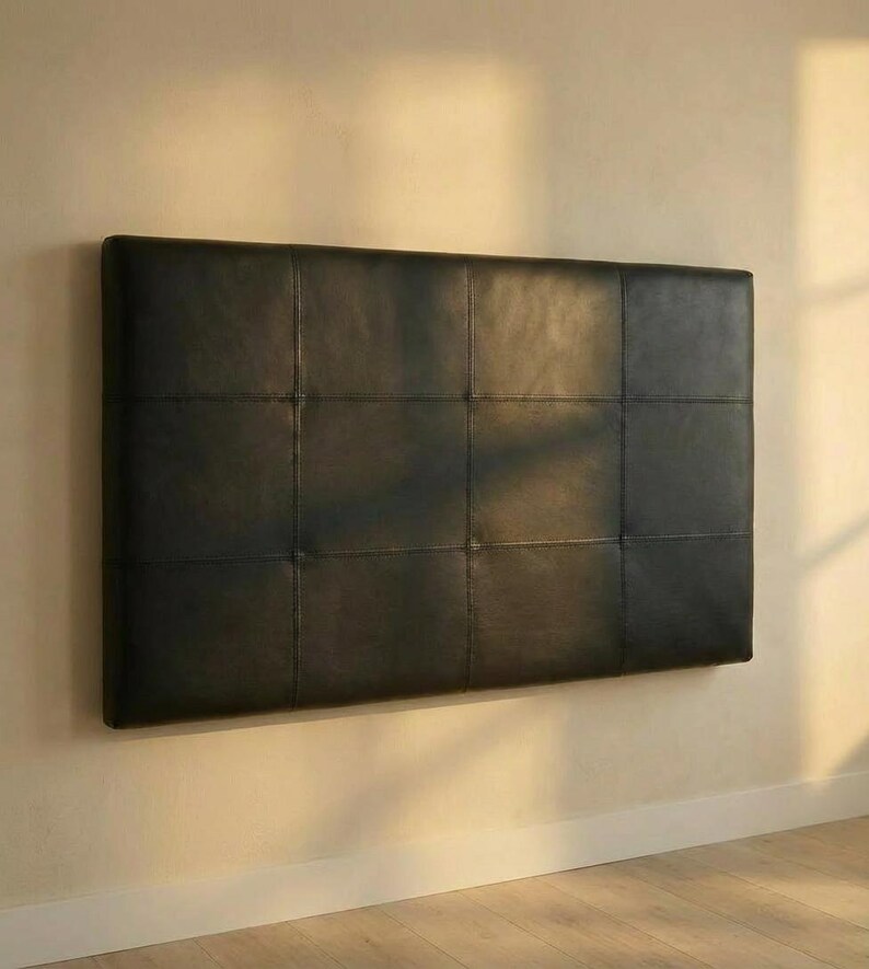 May include: A rectangular, black leather headboard with a grid pattern of stitched squares. The headboard is mounted on a light-colored wall, creating a minimalist aesthetic. The headboard is a piece of bedroom furniture.
