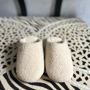 May include: A pair of white fluffy slippers with a soft, fuzzy texture. The slippers are on a black and white patterned surface.