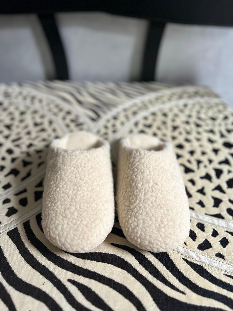 May include: A pair of white fluffy slippers with a soft, fuzzy texture. The slippers are on a black and white patterned surface.