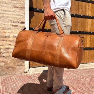 May include: A large, brown leather duffel bag with two top handles and a shoulder strap. The bag has a rectangular shape with a zipper closure and is held by a person. The bag is made of a distressed leather material.