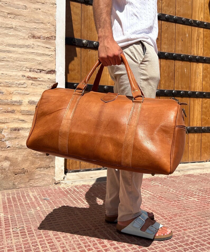 May include: A large, brown leather duffel bag with two top handles and a shoulder strap. The bag has a rectangular shape with a zipper closure and is held by a person. The bag is made of a distressed leather material.
