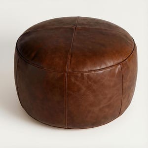 May include: A round, brown leather ottoman. The ottoman is a rich, dark brown color and has a stitched design on the top. The leather appears to be smooth and has a slight sheen. The ottoman is a solid, cylindrical shape.