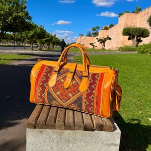 May include: A large, orange leather duffel bag with a patterned textile center panel. The textile features geometric designs in red, brown, and gold. The bag has a top handle, side pocket, and buckle closure. It is sitting on a wooden bench.