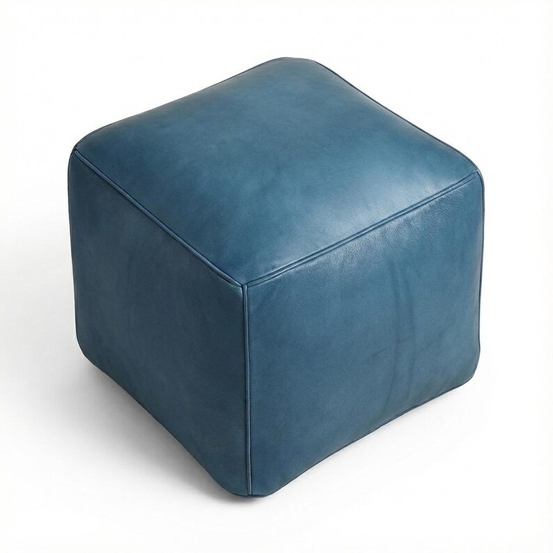 May include: A square, blue leather ottoman. The cube-shaped furniture piece has rounded edges and visible stitching along the seams. The ottoman is a solid teal color and sits on a white background.
