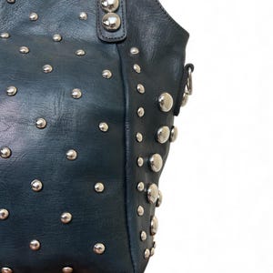 May include: Dark teal leather handbag with silver-tone studs. The bag features a rounded top, a handle, and a shoulder strap attachment. The studs are evenly spaced across the surface of the bag, adding a decorative element.