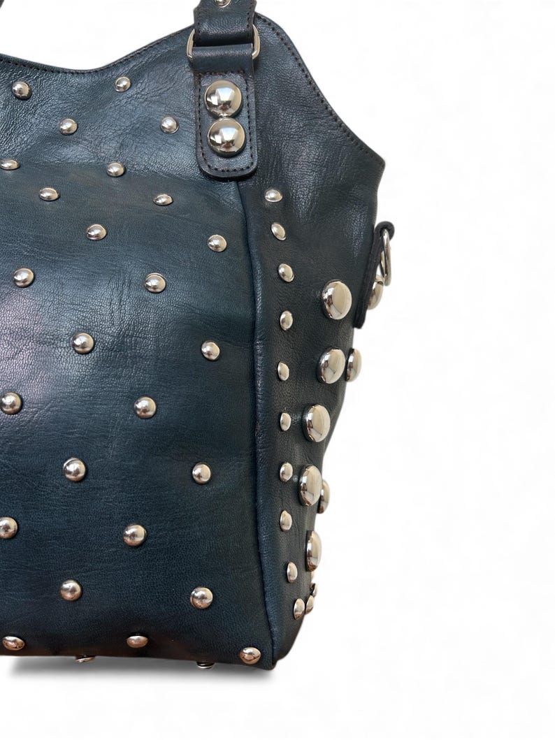 May include: Dark teal leather handbag with silver-tone studs. The bag features a rounded top, a handle, and a shoulder strap attachment. The studs are evenly spaced across the surface of the bag, adding a decorative element.