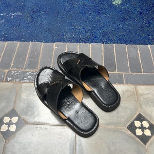 May include: A pair of black leather sandals with crisscross straps. The sandals have a dark sole and are placed on a gray tile surface. The background features a blue pool.