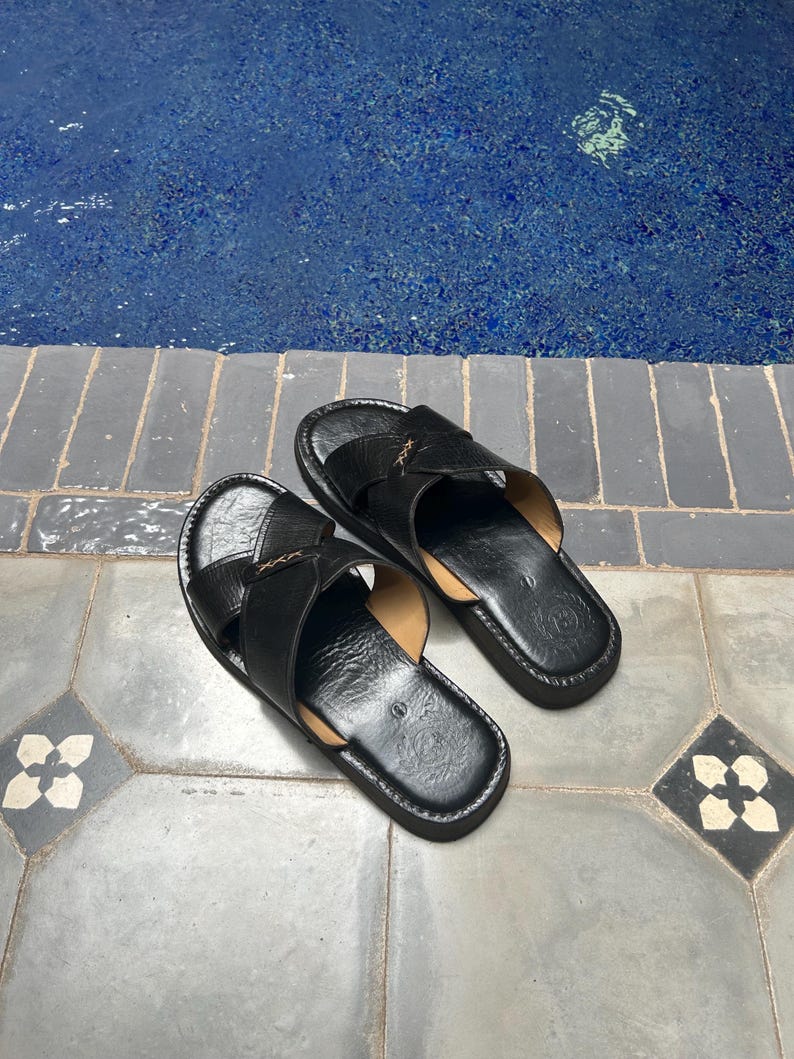 May include: A pair of black leather sandals with crisscross straps. The sandals have a dark sole and are placed on a gray tile surface. The background features a blue pool.