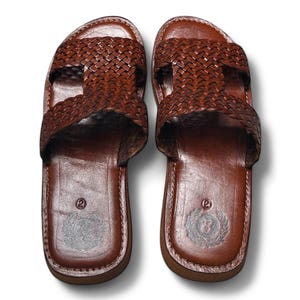 May include: A pair of brown leather sandals. The sandals feature a woven strap design across the top and a smooth leather footbed. The soles are a darker brown, and the sandals appear to be new.