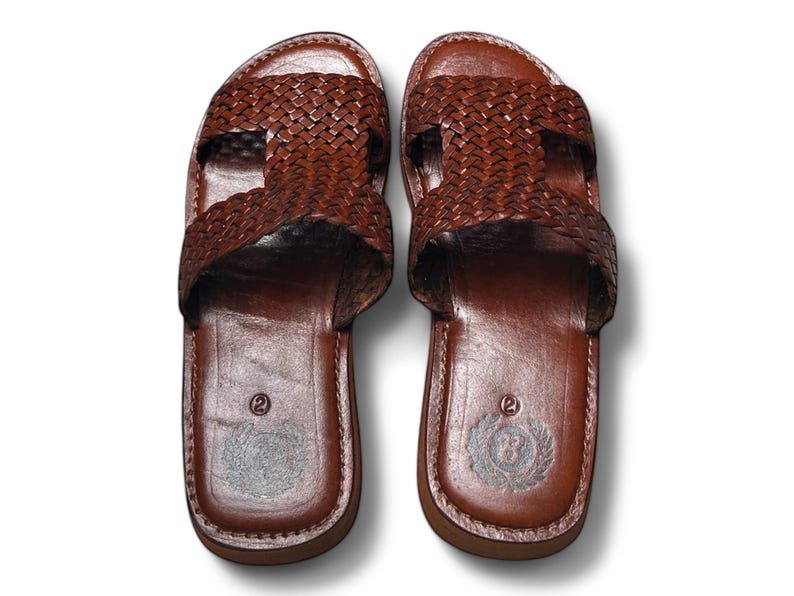 May include: A pair of brown leather sandals. The sandals feature a woven strap design across the top and a smooth leather footbed. The soles are a darker brown, and the sandals appear to be new.