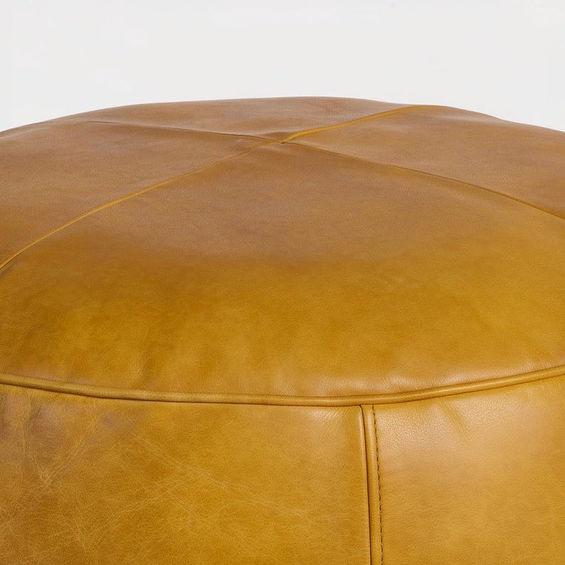 May include: Close-up of a round, mustard-colored leather ottoman. The ottoman has a stitched design on the top and a seam around the base. The leather has a slightly distressed appearance.