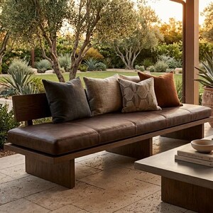 K&ouml;nnte beinhalten: Eine moderne Outdoor-Bank mit dunkelbraunem Lederkissen und Holzrahmen. Die Bank ist mit mehreren dekorativen Kissen in Grau-, Beige- und Rostt&ouml;nen geschm&uuml;ckt. Die Szene spielt in einem gepflegten Garten mit &uuml;ppigem Gr&uuml;n.