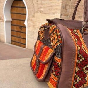May include: A colorful duffel bag with a patterned fabric design in shades of red, orange, and yellow. The bag features brown leather accents, including the handles and a small side pocket with a buckle. The bag is sitting on a stone surface.