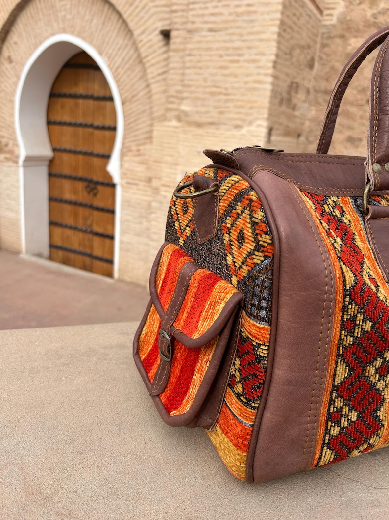 May include: A colorful duffel bag with a patterned fabric design in shades of red, orange, and yellow. The bag features brown leather accents, including the handles and a small side pocket with a buckle. The bag is sitting on a stone surface.