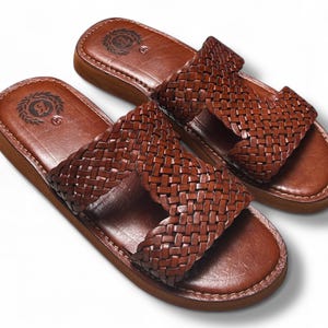 May include: A pair of brown leather sandals with a woven strap design. The sandals have a light brown sole and a dark brown leather footbed. The sandals are displayed on a white background.