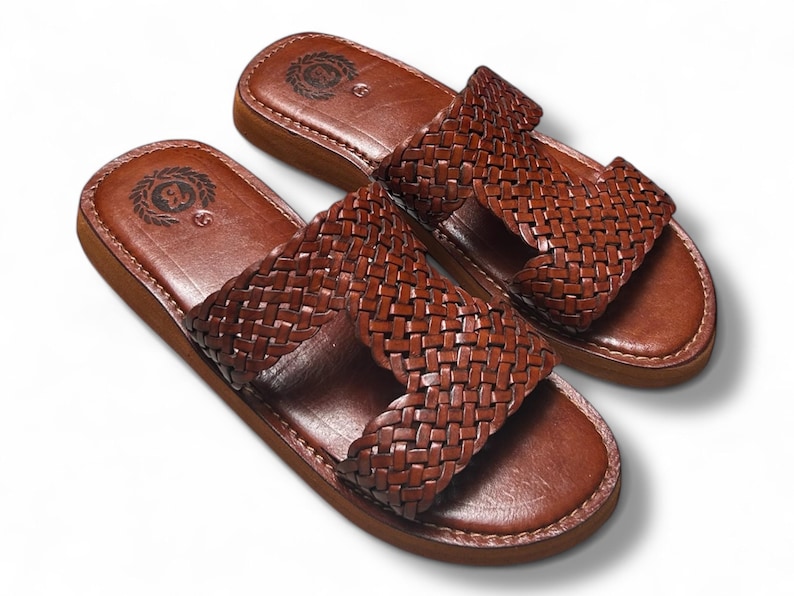 May include: A pair of brown leather sandals with a woven strap design. The sandals have a light brown sole and a dark brown leather footbed. The sandals are displayed on a white background.