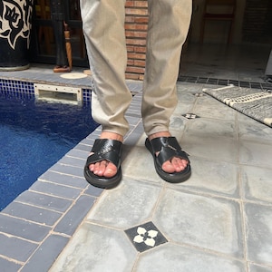 May include: Black leather sandals with a crisscross strap design. The sandals have a flat sole and are worn with light-colored pants. The image is taken outdoors, with a pool and decorative tile in the background.
