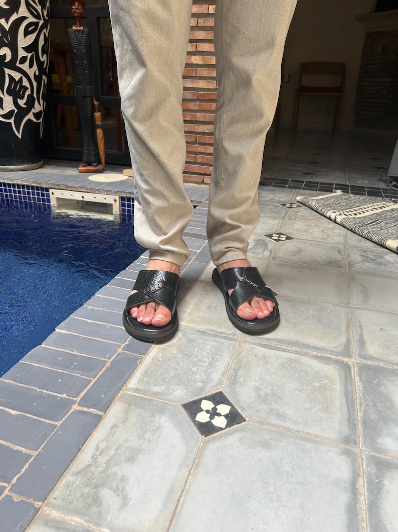May include: Black leather sandals with a crisscross strap design. The sandals have a flat sole and are worn with light-colored pants. The image is taken outdoors, with a pool and decorative tile in the background.