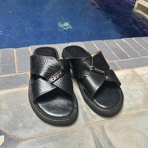 May include: A pair of black leather sandals with a cross-strap design. The sandals have a textured appearance and a dark sole. The image shows the sandals on a tiled surface, with a pool in the background.