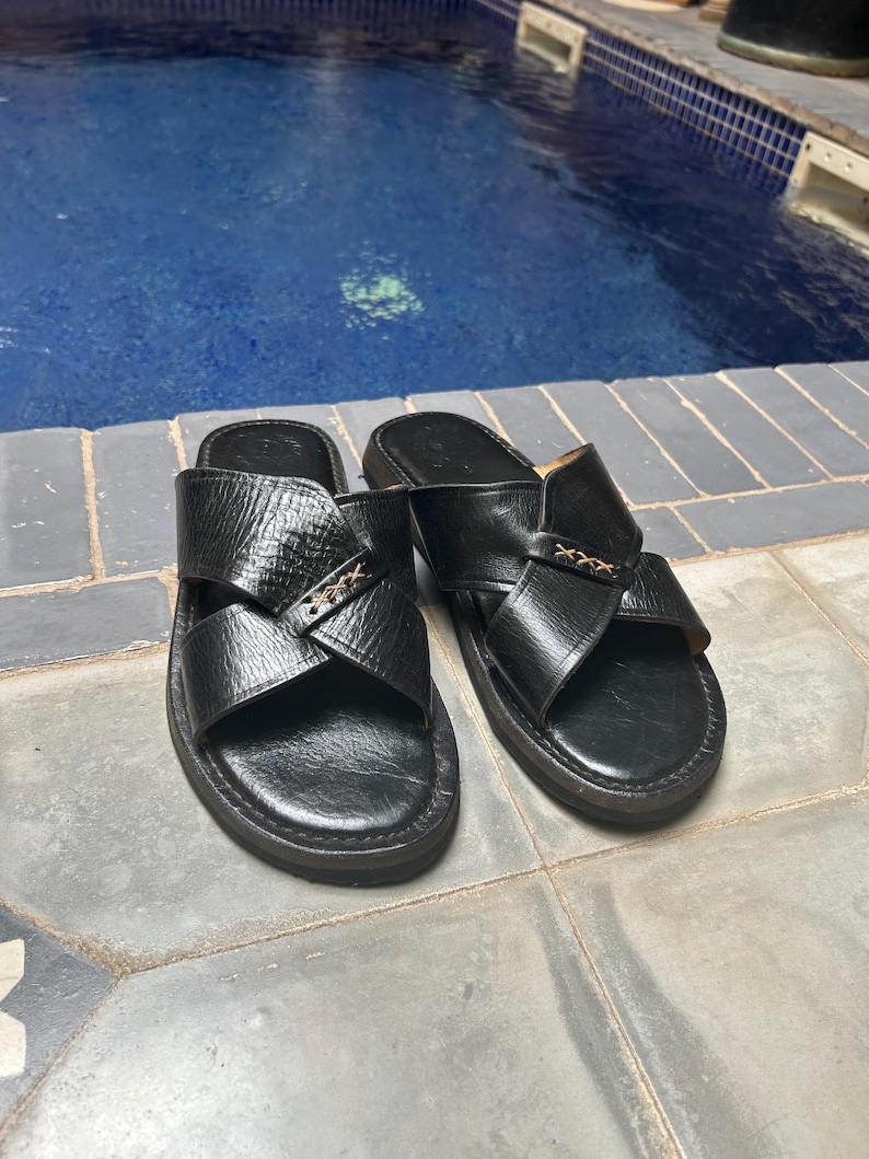 May include: A pair of black leather sandals with a cross-strap design. The sandals have a textured appearance and a dark sole. The image shows the sandals on a tiled surface, with a pool in the background.