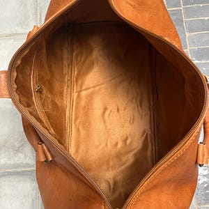 May include: Open, empty, brown leather duffel bag with a zipper closure. The bag has a rectangular shape with rounded ends and a lighter brown interior. The zipper is gold-colored, and the bag has two small handles on each side.