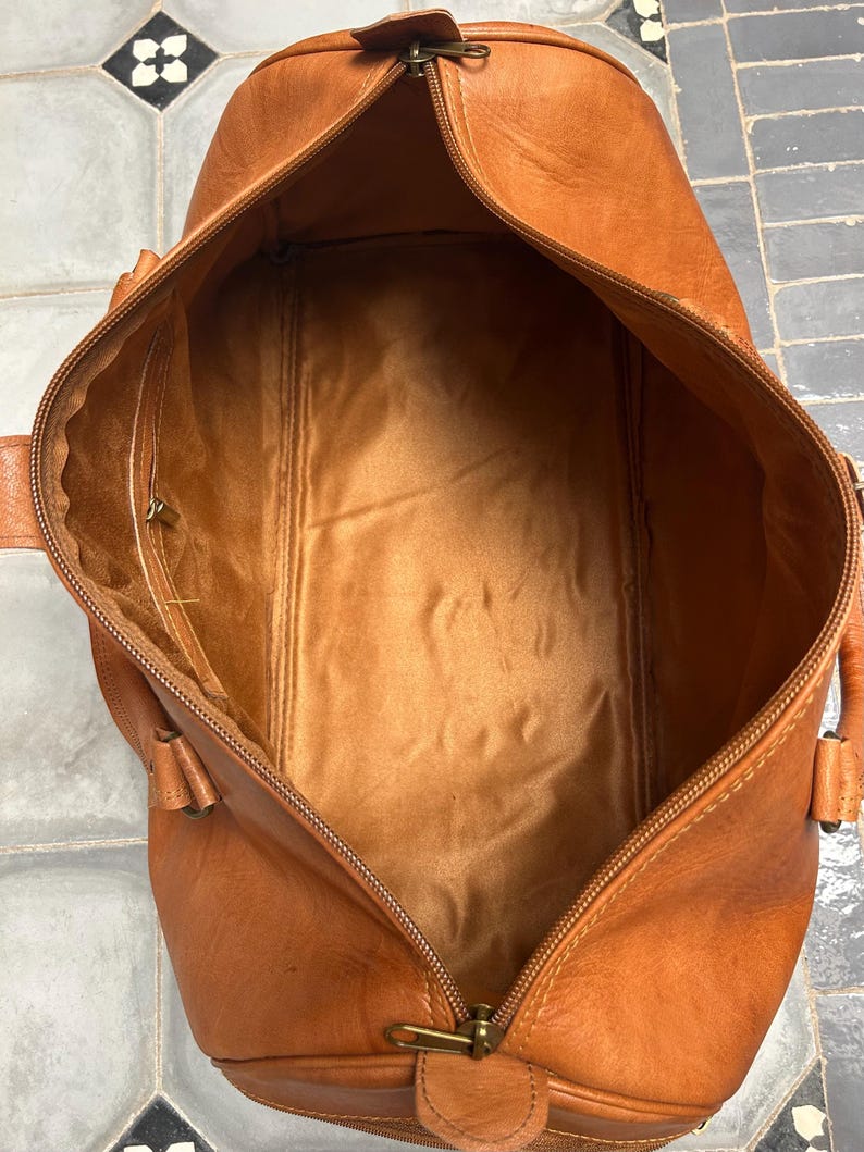 May include: Open, empty, brown leather duffel bag with a zipper closure. The bag has a rectangular shape with rounded ends and a lighter brown interior. The zipper is gold-colored, and the bag has two small handles on each side.
