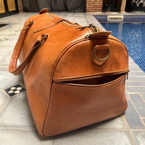 May include: A large, brown leather duffel bag with a zippered pocket and handles. The bag has a rounded shape and a brass zipper. The bag is made of a smooth, high-quality leather. The bag is designed for travel and carrying luggage.