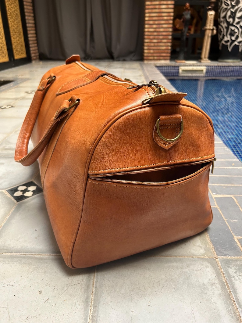 May include: A large, brown leather duffel bag with a zippered pocket and handles. The bag has a rounded shape and a brass zipper. The bag is made of a smooth, high-quality leather. The bag is designed for travel and carrying luggage.