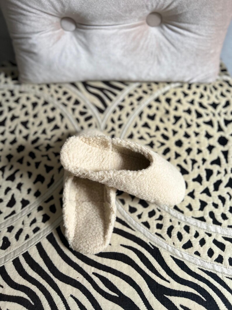 May include: A pair of white fluffy slippers with a soft, fuzzy texture. The slippers are on a black and white patterned surface.