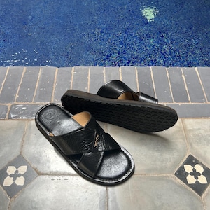 May include: A pair of black leather sandals with a criss-cross design on the top. The sandals have a dark brown sole and are placed on a patterned tile surface near a pool of water.