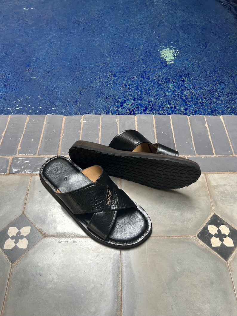 May include: A pair of black leather sandals with a criss-cross design on the top. The sandals have a dark brown sole and are placed on a patterned tile surface near a pool of water.